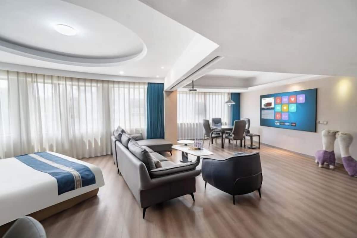 Quarto de hotel moderno com cama, sofá, poltrona, mesa de jantar e televisão fixada na parede.