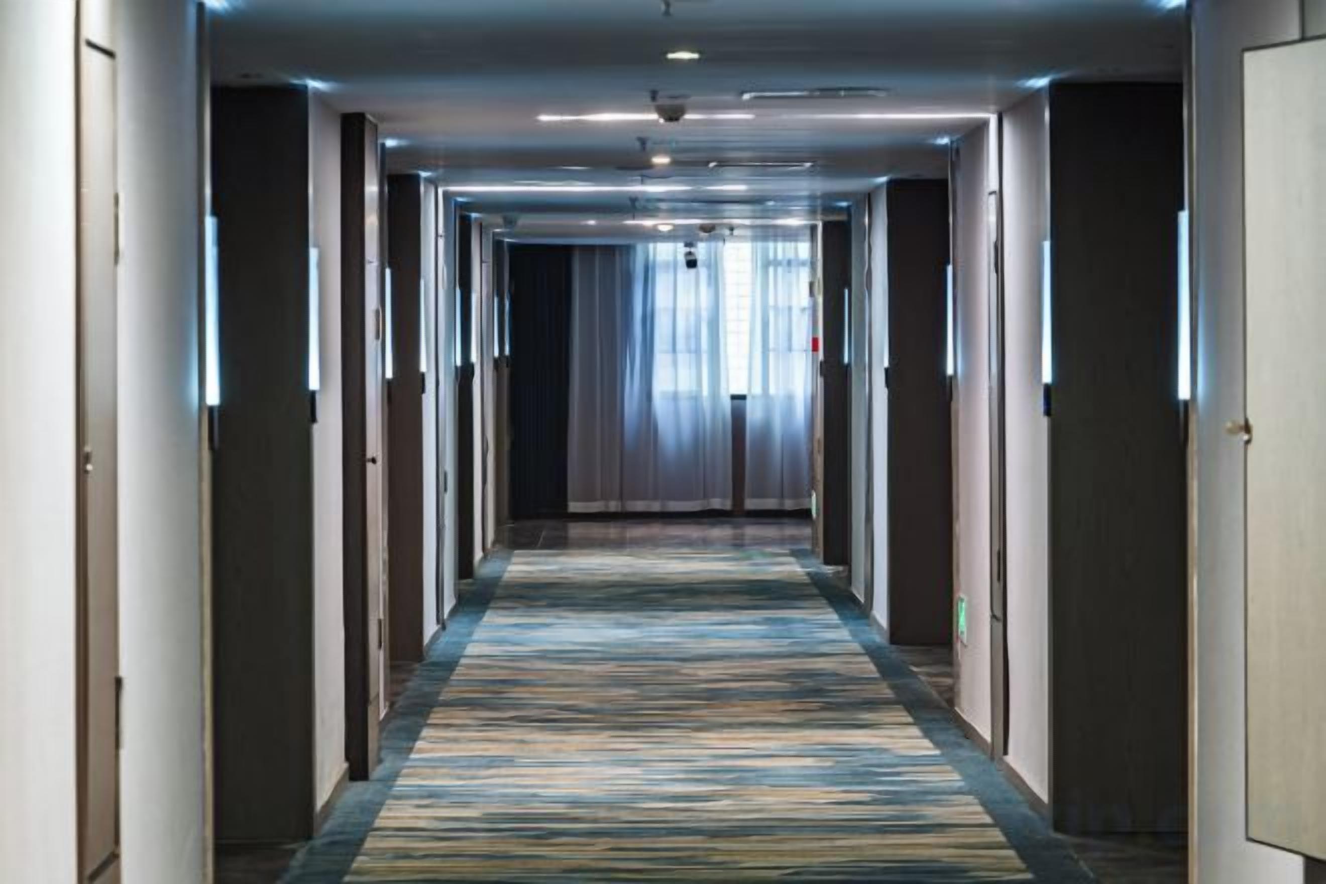 Un couloir d'hôtel avec un sol recouvert de moquette, des portes de chaque côté et un luminaire au plafond.