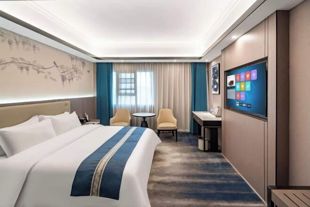 Quarto de hotel com uma cama grande, uma escrivaninha, duas cadeiras, uma televisão e uma janela com cortinas.