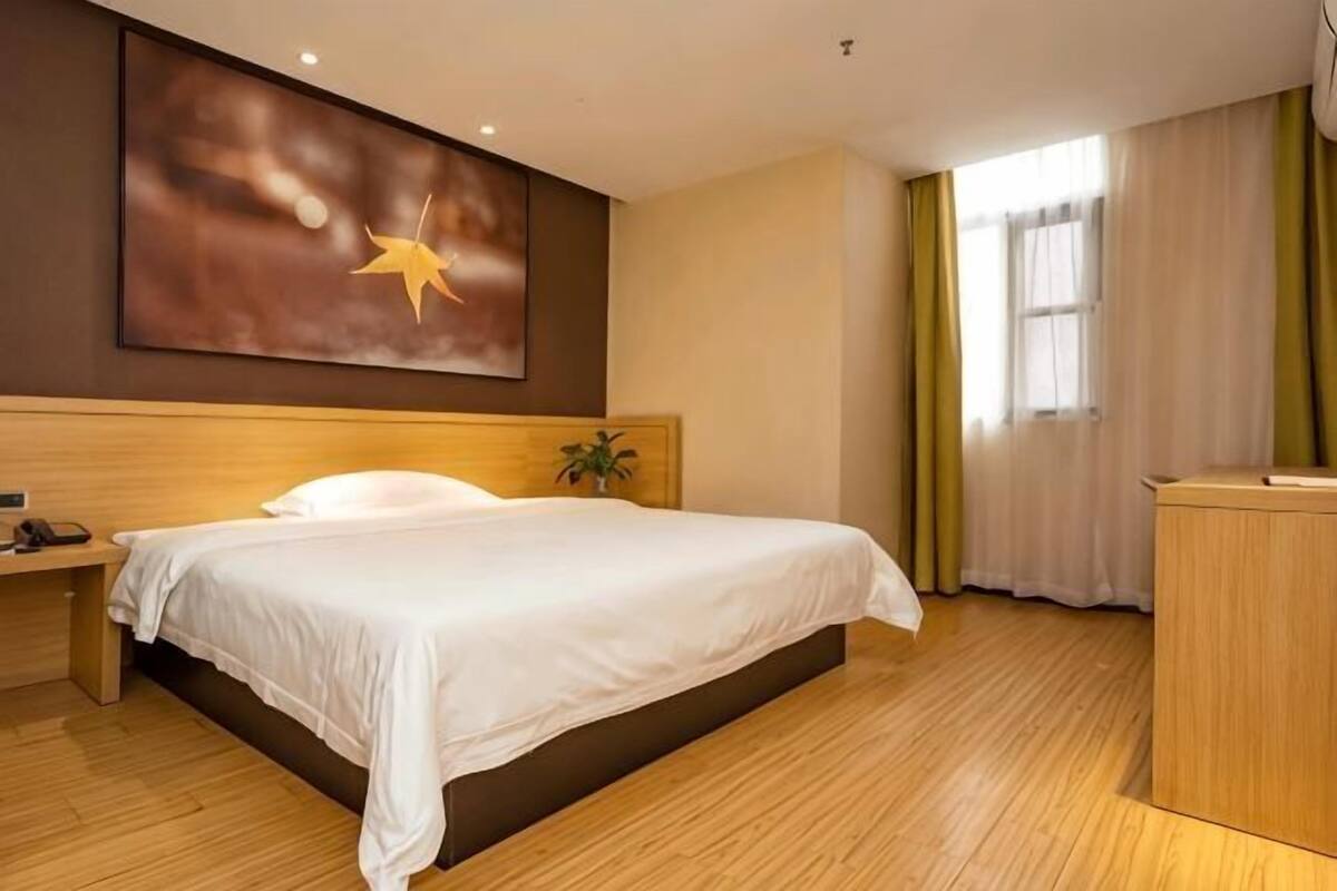 Hotellihuone, jossa on suuri sänky, puinen pääty, työpöytä tuolilla, yöpöytä, suuri ikkuna verhoilla ja seinälle kiinnitetty taideteos.