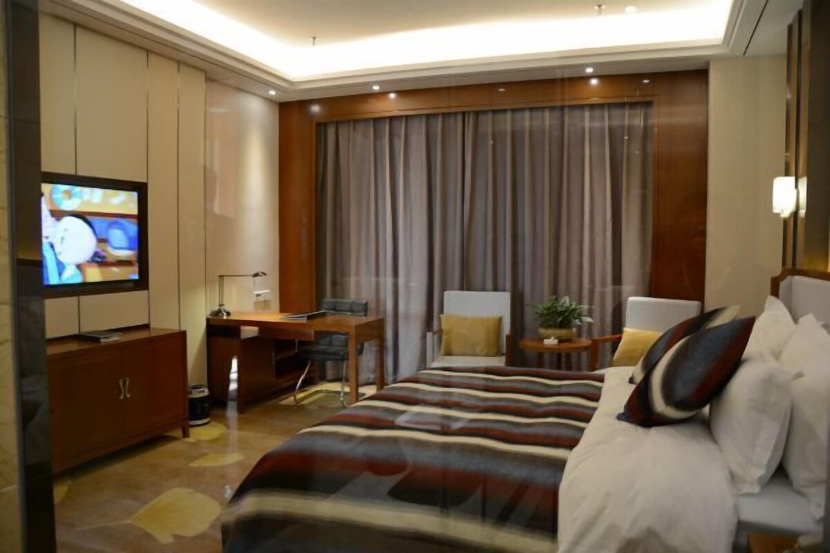 A modern hotel room with a large bed, a desk, a television, and a large window with curtains.