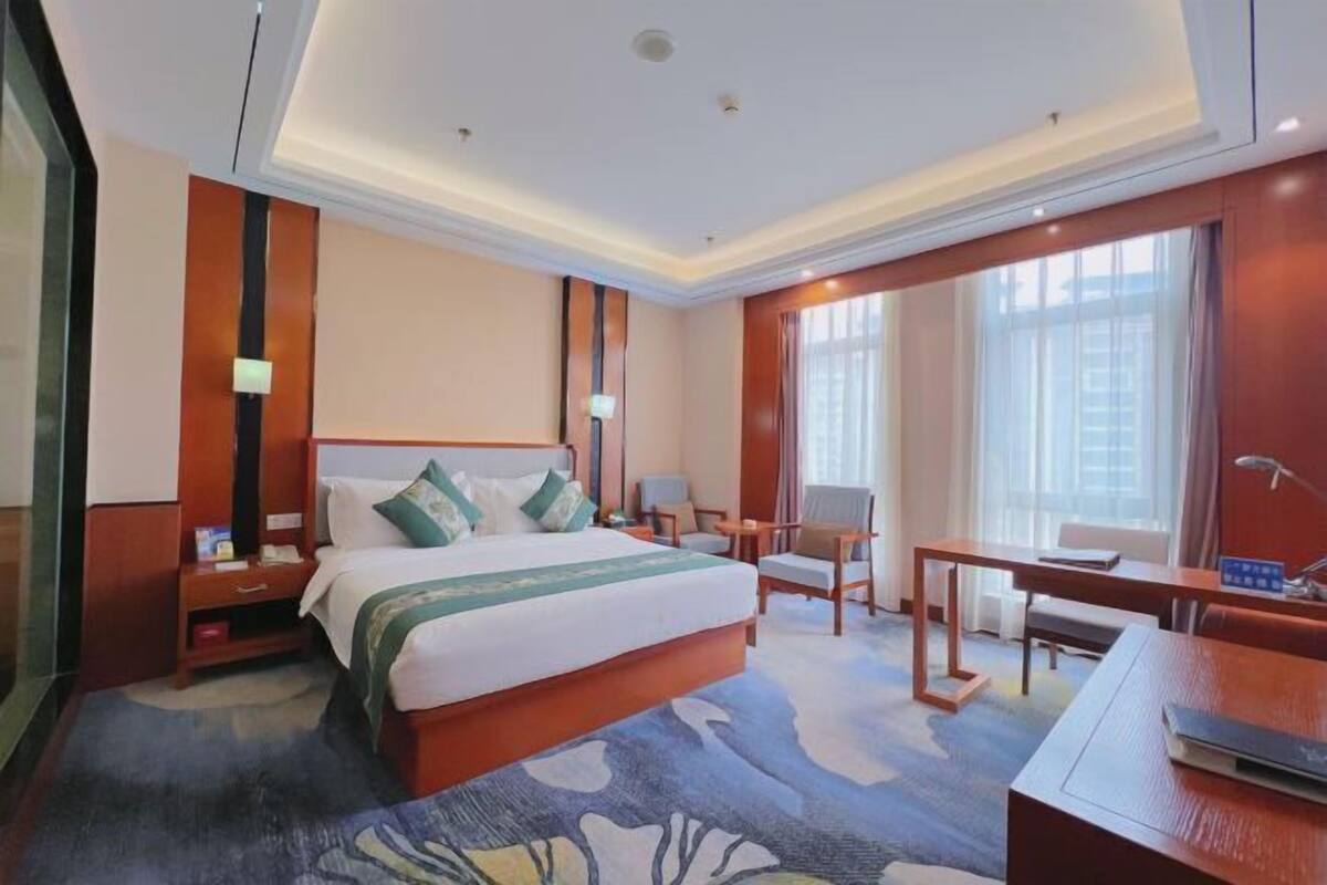 A hotel room with a large bed, a desk, two chairs, a nightstand, and a window with curtains.