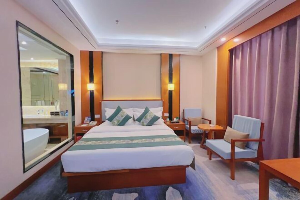 A hotel room with a large bed, bedside tables, a chair, a desk, a mirror, and a bathroom with a sink and toilet.