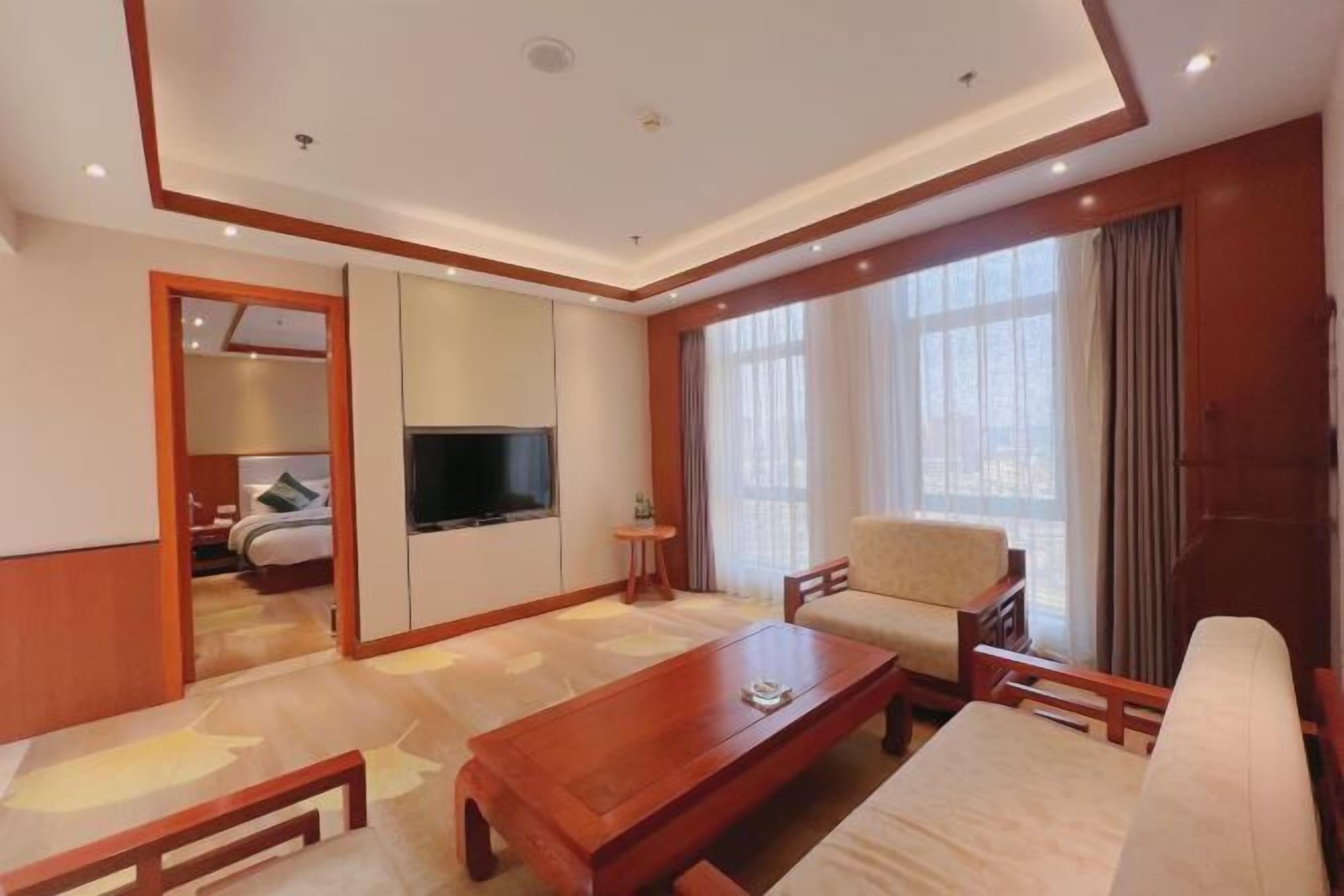 Habitación de hotel con una mesa de centro de madera, un sofá, un televisor y un ventanal con cortinas.