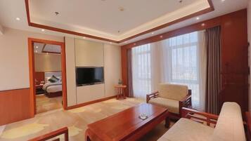 Habitación de hotel con una mesa de centro de madera, un sofá, un televisor y un ventanal con cortinas.