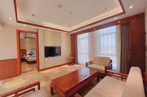 Habitación de hotel con una mesa de centro de madera, un sofá, un televisor y un ventanal con cortinas.