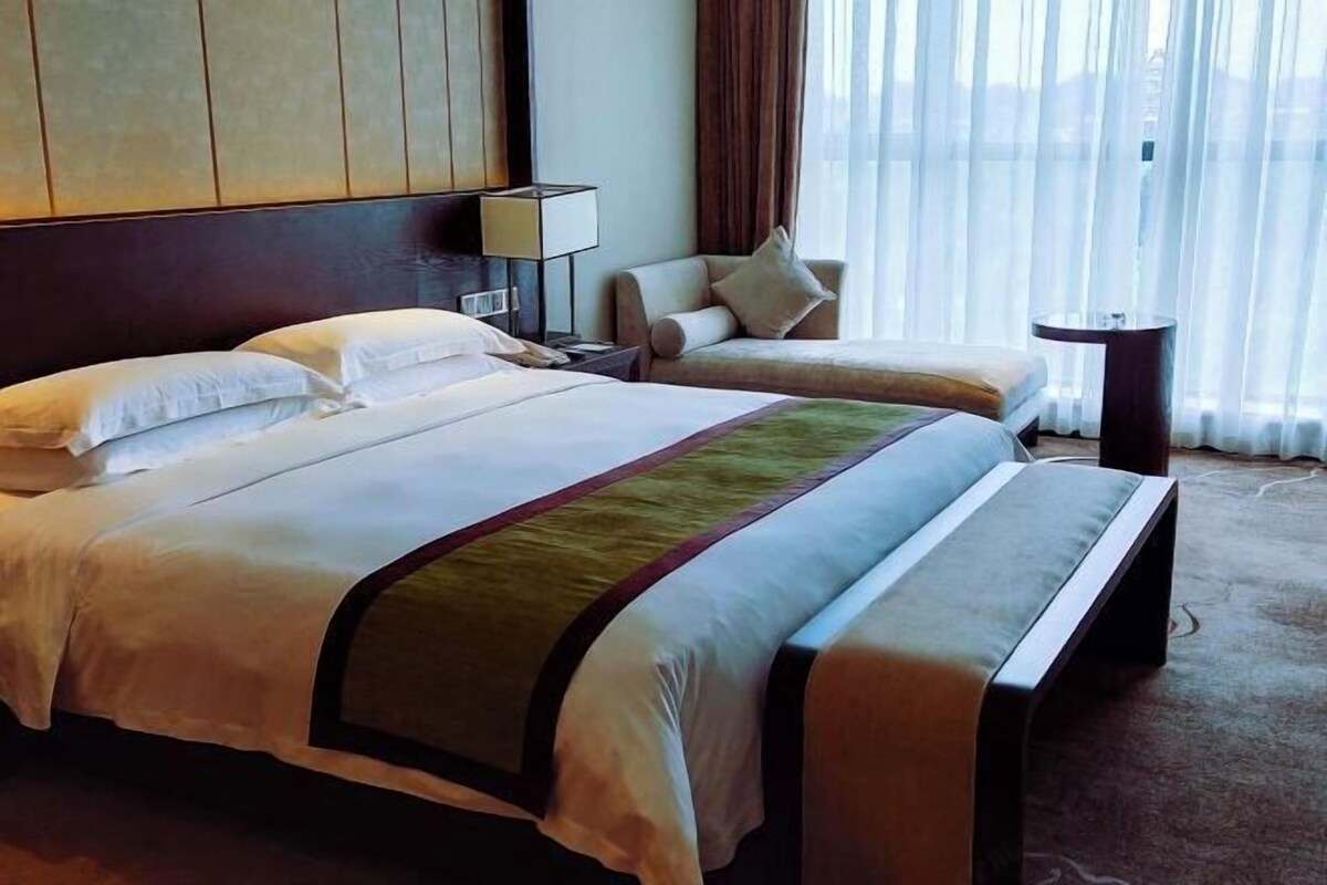 A hotel room with a large bed, a smaller bed, a nightstand, a lamp, a chair, and a view of the outdoors through sheer curtains.