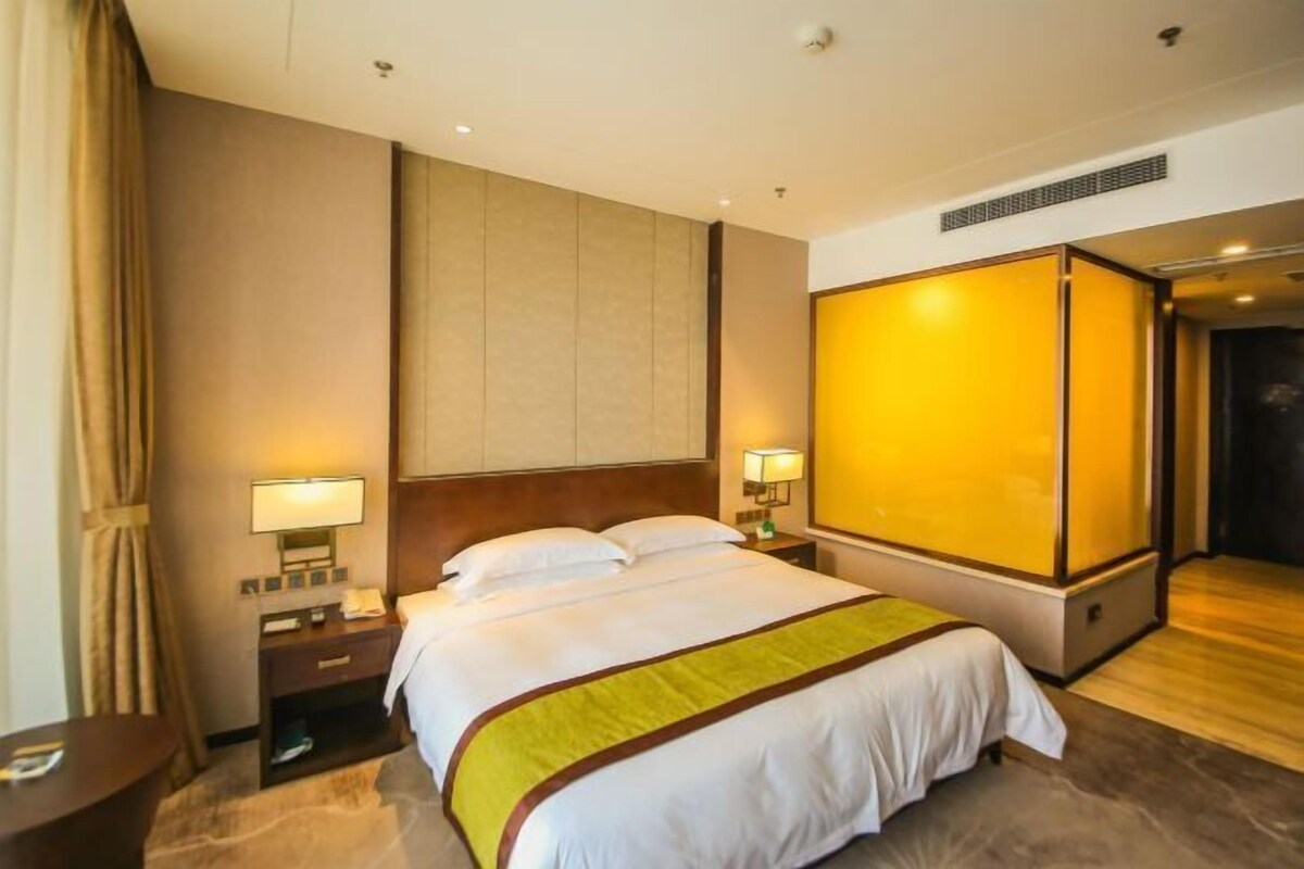 A hotel room with a bed, bedside tables, lamps, a large window, and a door leading to another room.