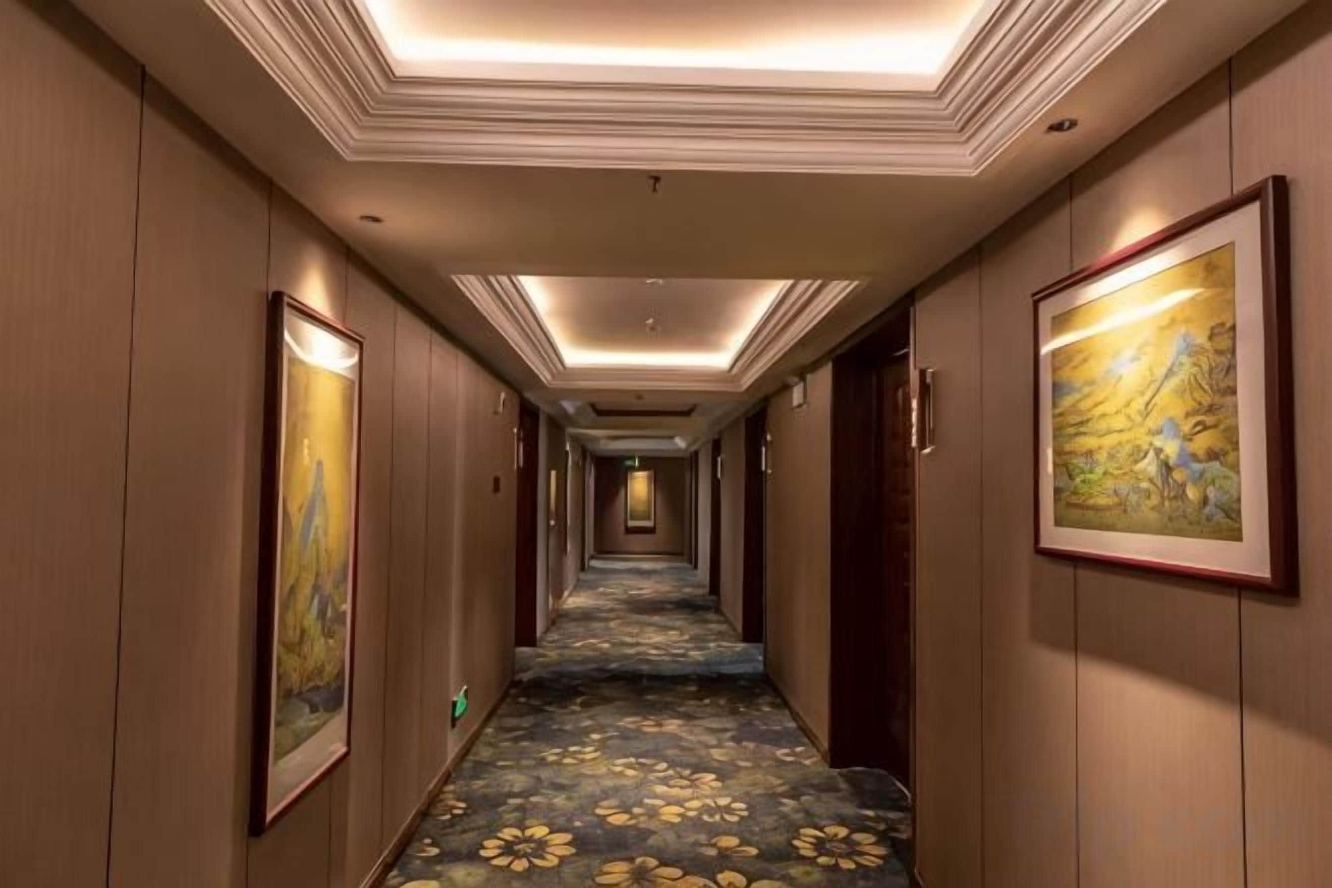 A hotel hallway with a carpeted floor, framed artwork on the walls, and a ceiling with recessed lighting.