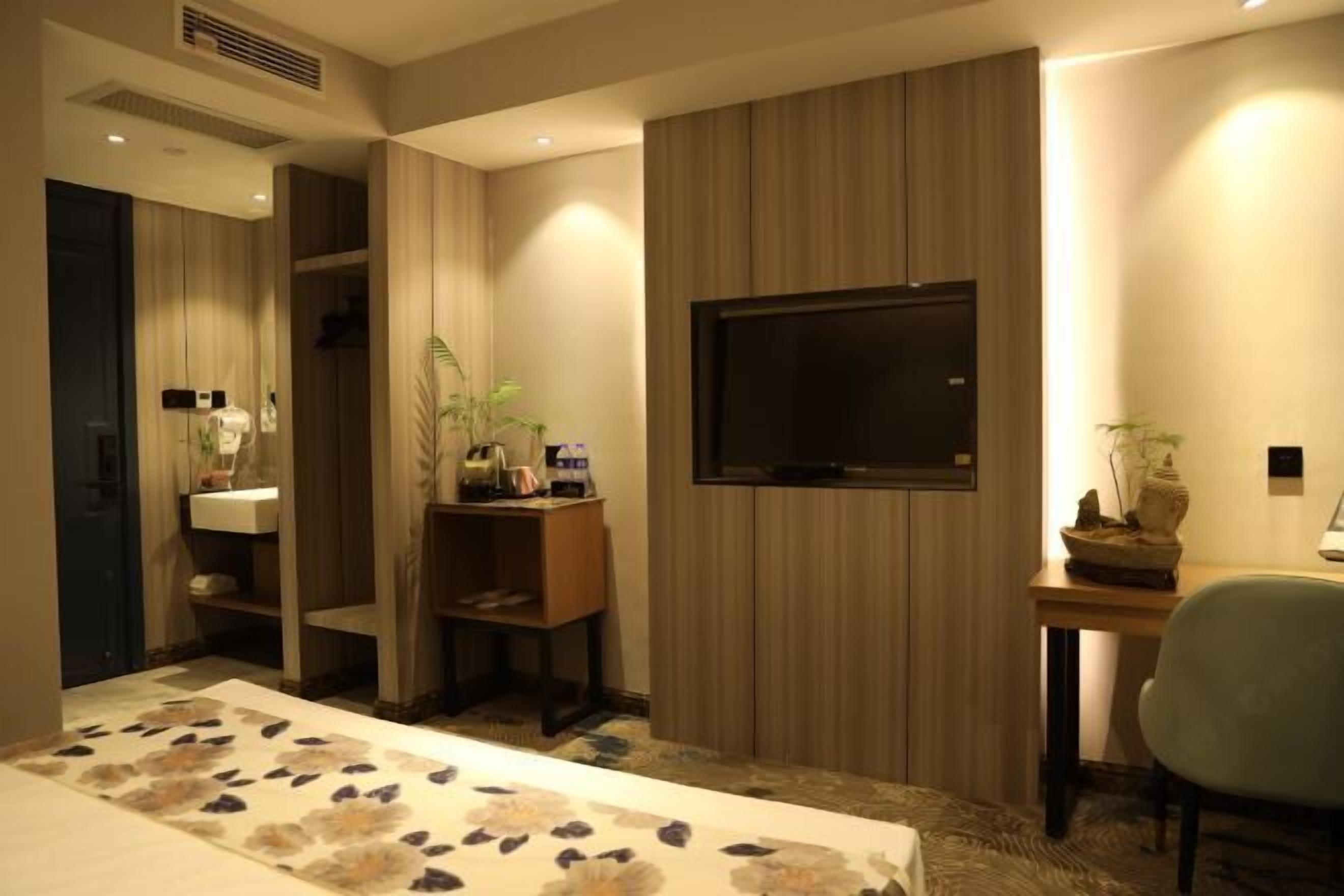 Una camera d'albergo con un letto, una scrivania con televisore, una poltrona, una pianta e uno scaffale con articoli da toilette.