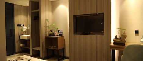 A hotel room with a bed, a desk with a TV, a chair, a plant, and a shelf with toiletries.