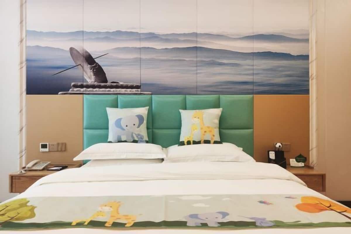 Een hotelkamer met een bed, voorzien van een berglandschap op de muur, een groen gestoffeerd hoofdeinde en een deken met dierenprint.