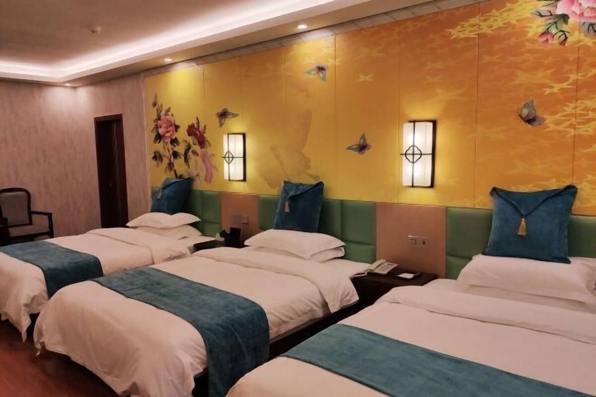 A hotel room with three beds, each with a blue and white bedspread. The room features a yellow wall with floral and butterfly decorations, and two wall-mounted lamps.