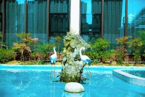Una fuente con dos estatuas blancas en un estanque, rodeada de vegetación y un edificio moderno al fondo.