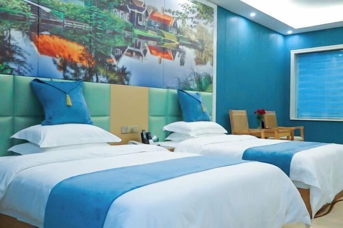 Een hotelkamer met twee bedden, een groot raam en een muurschildering van een dorpsgezicht.