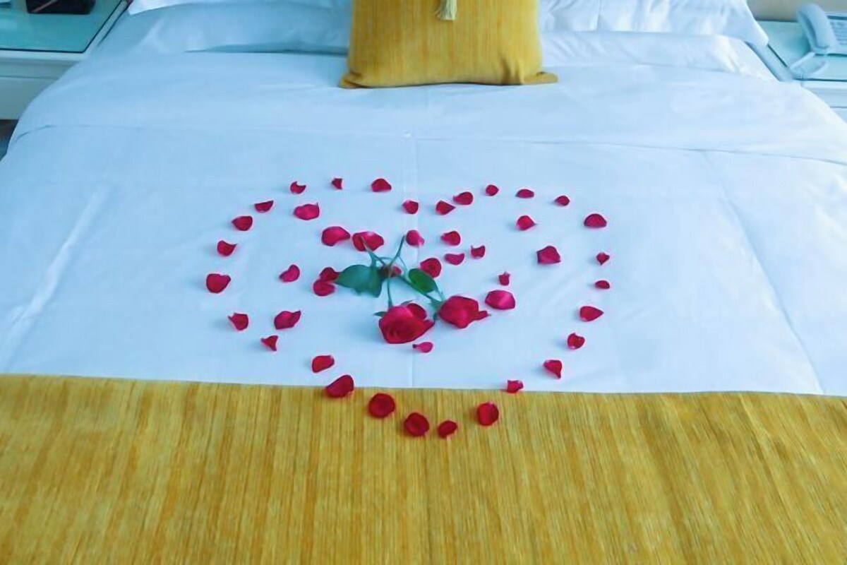 A bed with a white bedspread, a yellow pillow, and a heart-shaped arrangement of red rose petals and a single red rose.