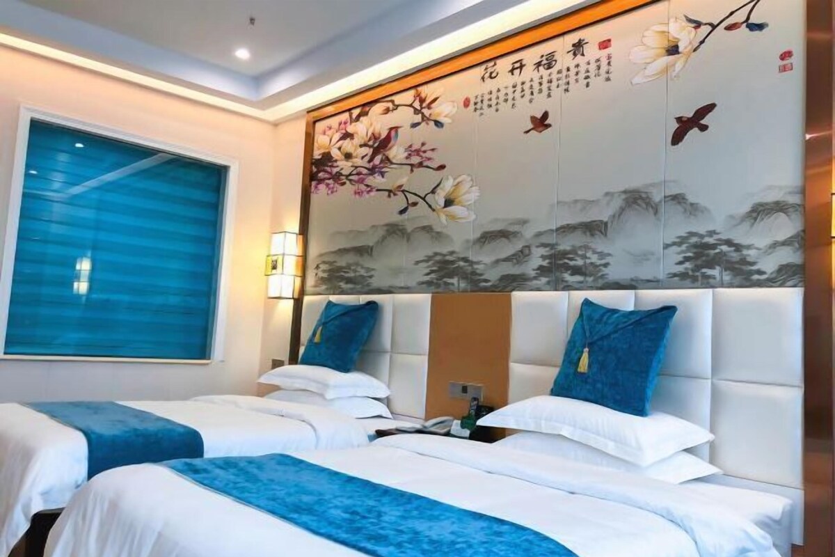 A hotel room with two beds, a large window with blue blinds, and a decorative wall with floral and bird paintings.