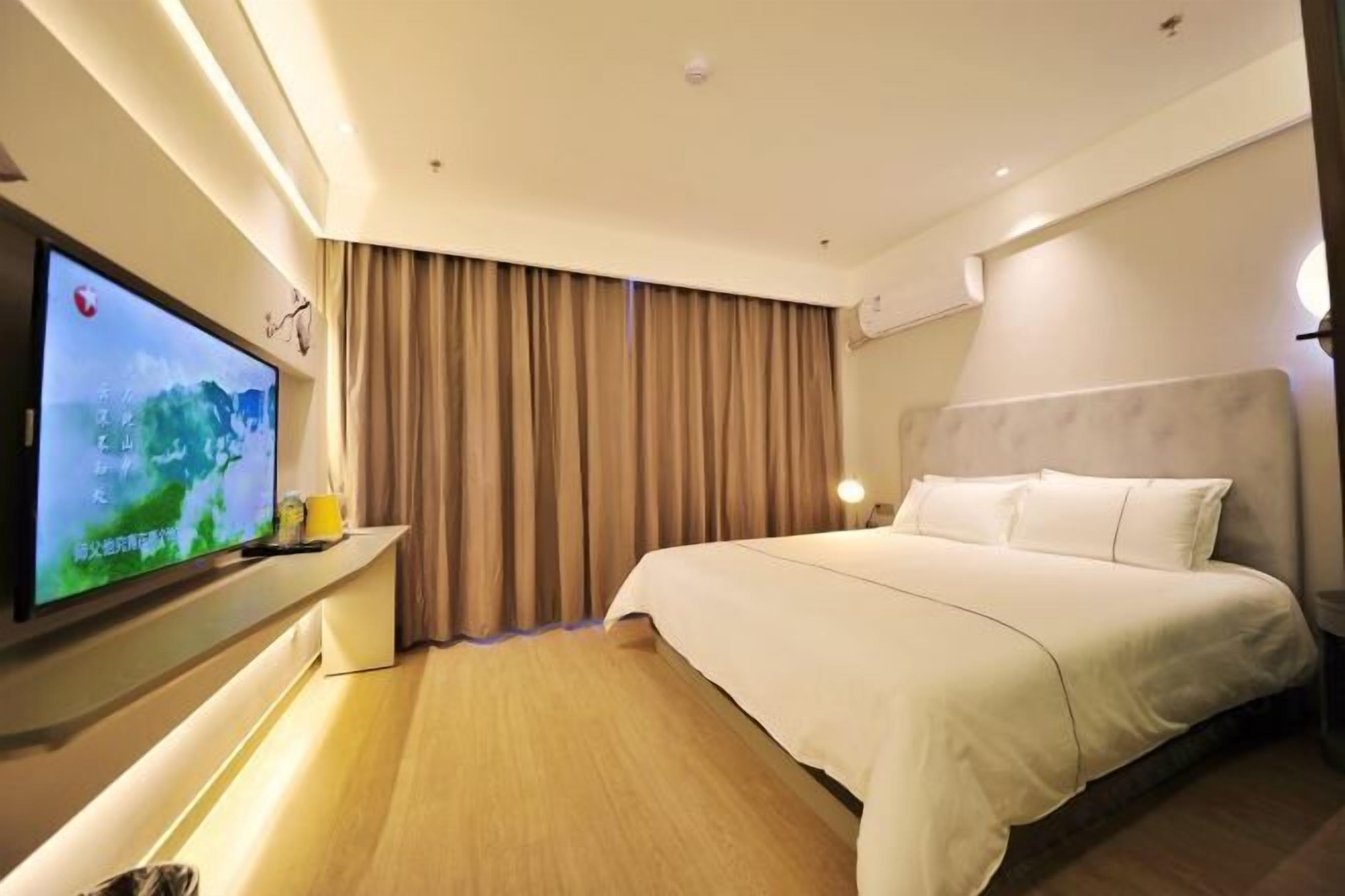 A hotel room with a large bed, a flat-screen TV mounted on the wall, a bedside table, and a window with curtains.