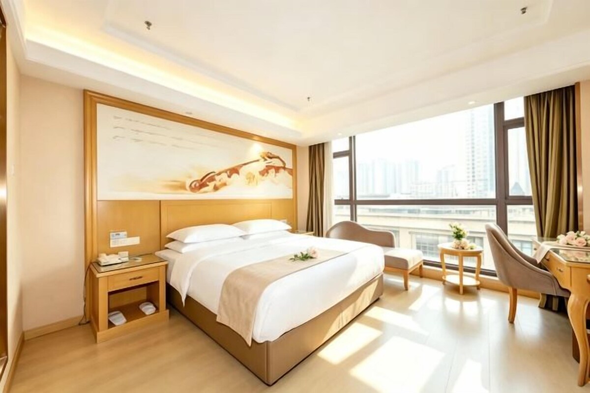 Habitación de hotel con una cama grande, mesita de noche, silla, escritorio y vistas a la ciudad.