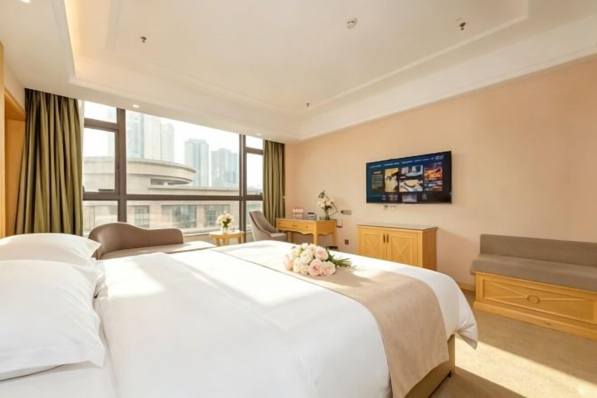Quarto de hotel com cama grande, escrivaninha, cadeira e vista para a paisagem urbana.