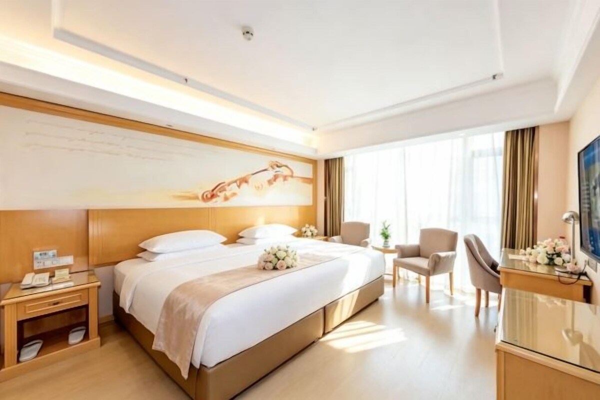A hotel room with a large bed, bedside tables, a desk with a chair, and a large window.