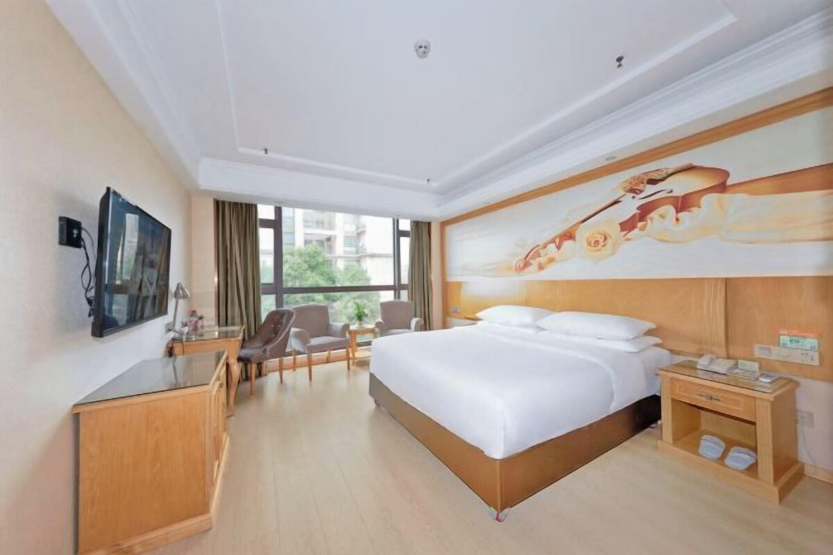 A hotel room with a bed, a desk, a chair, a TV, and a painting on the wall.