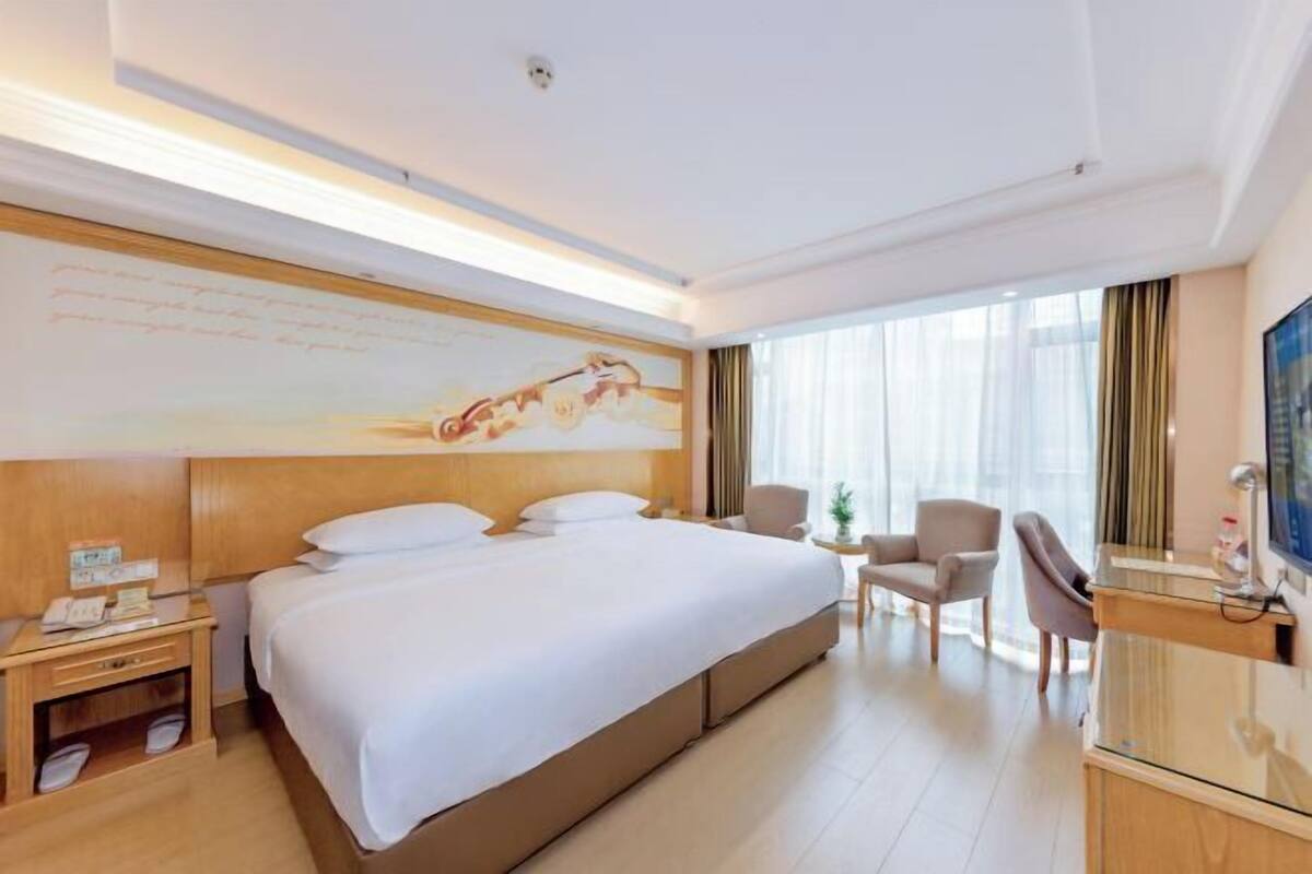 A hotel room with a large bed, a desk with a computer, a TV, and a window with curtains.