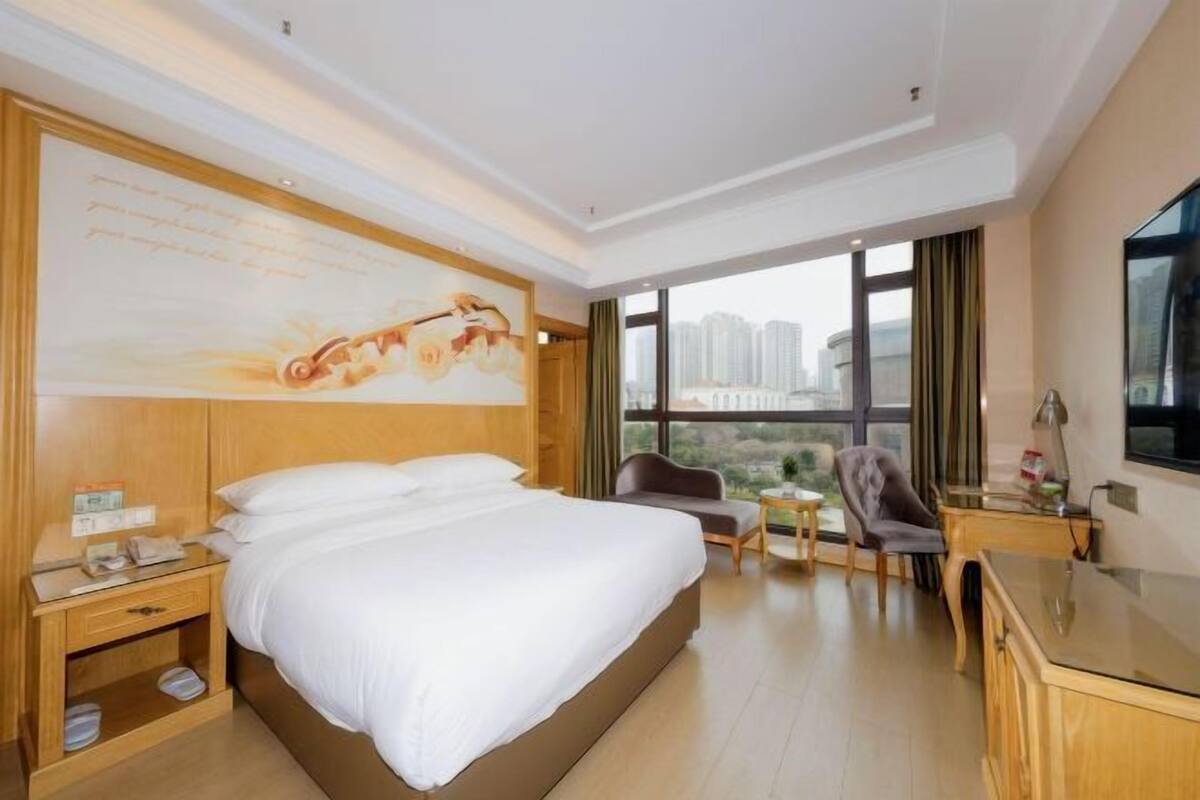 A hotel room with a large bed, a desk, a chair, a small table, and a large window offering a city view.