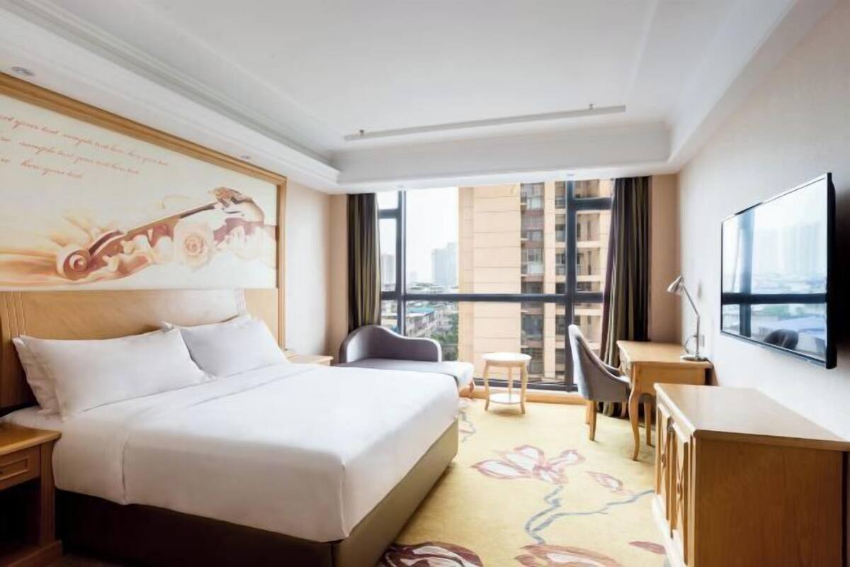 A hotel room with a large bed, a desk, a chair, a television, and a window with a city view.