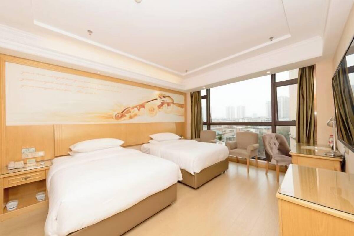 A hotel room with two beds, a desk, a chair, and a large window offering a city view.