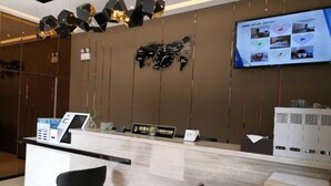 Zona de recepción de hotel con diseño moderno, que incluye un mostrador de recepción, dos sillas negras, una pantalla digital mostrando imágenes y un reloj de pared.