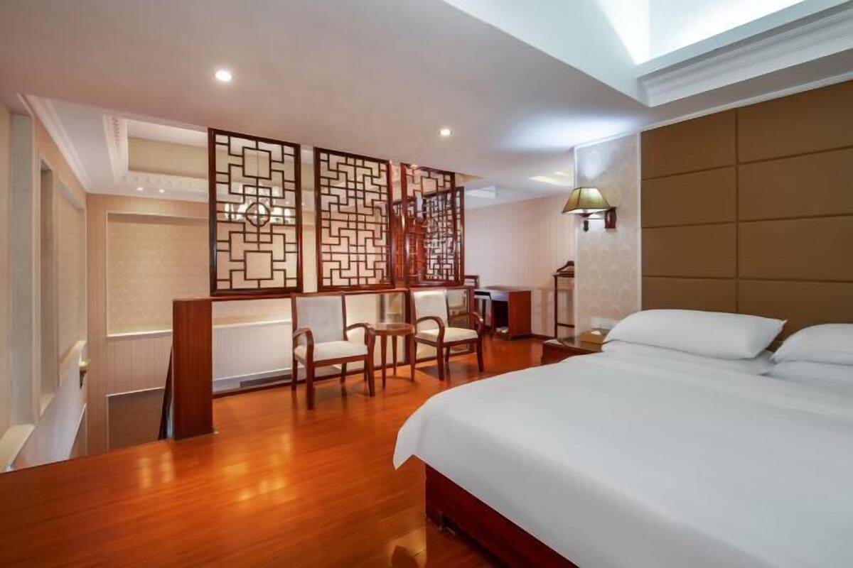 A hotel room with a large bed, wooden floor, a seating area with chairs, a decorative screen, and a lamp.