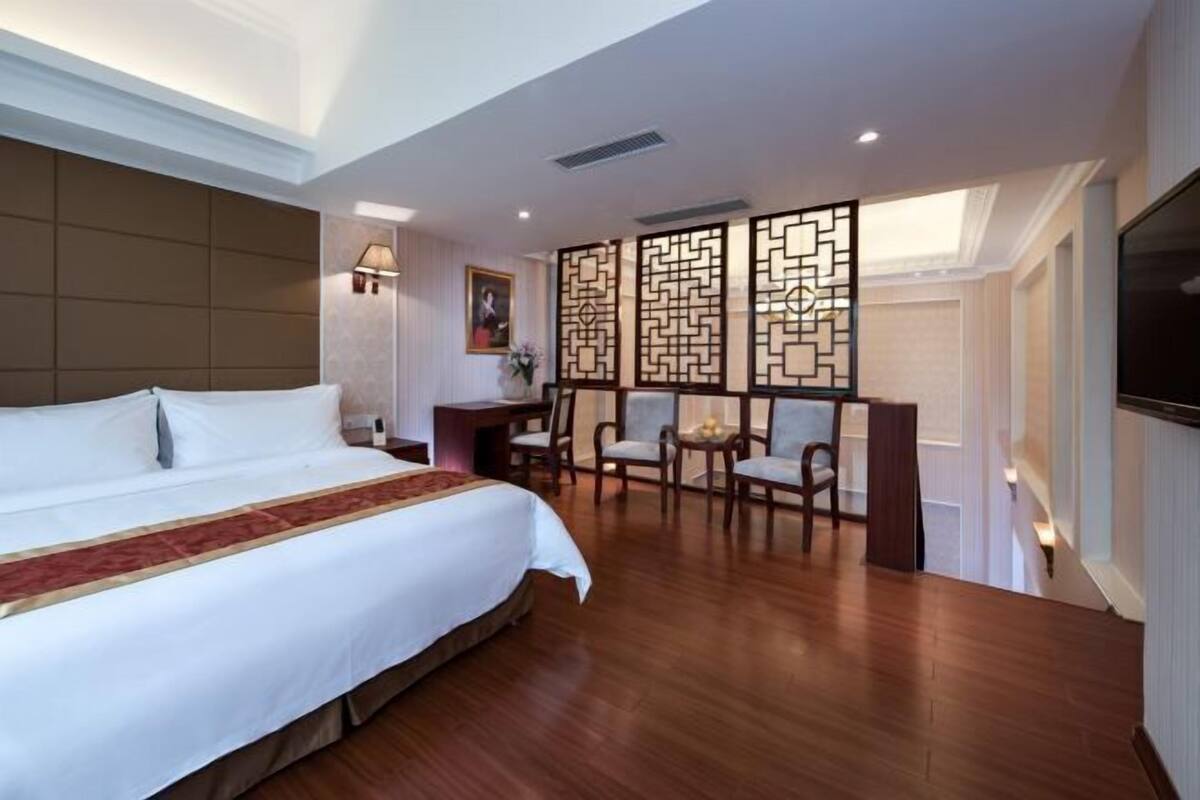 A hotel room with a large bed, a desk with chairs, a TV, and decorative screens.