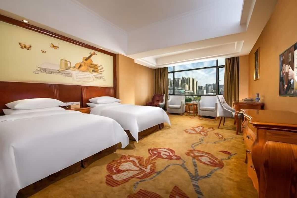 A hotel room with two beds, a city view, a floral carpet, and a desk with a chair.