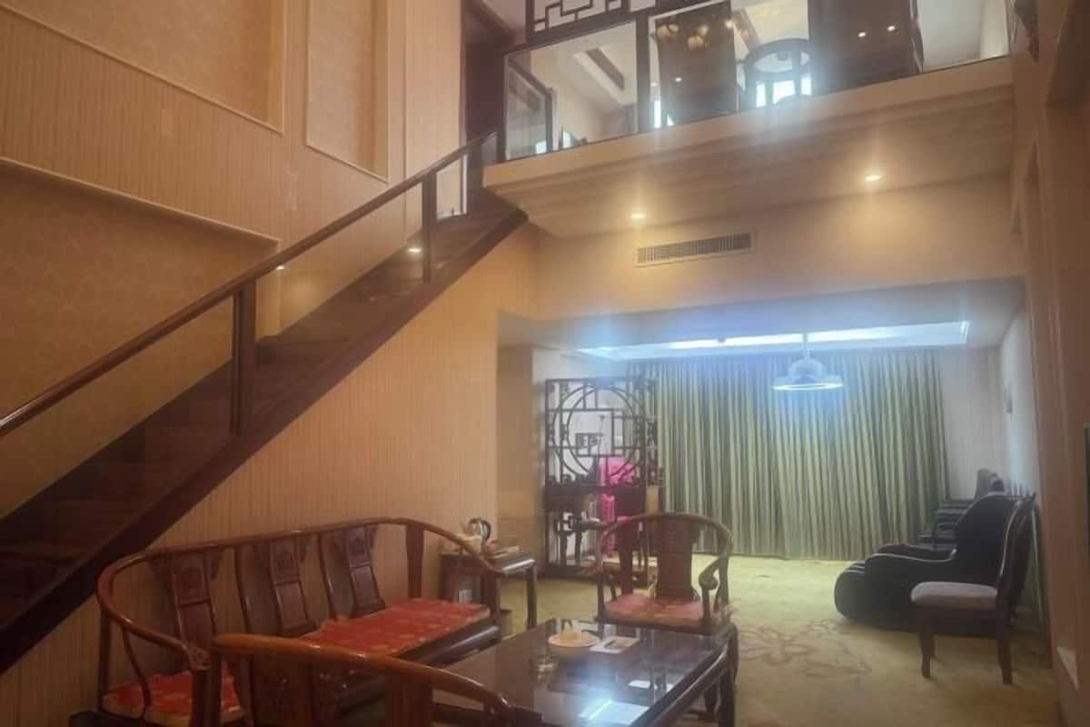 A high-ceilinged room with a wooden staircase, a dining area with wooden chairs, a sofa, and a large window with curtains.