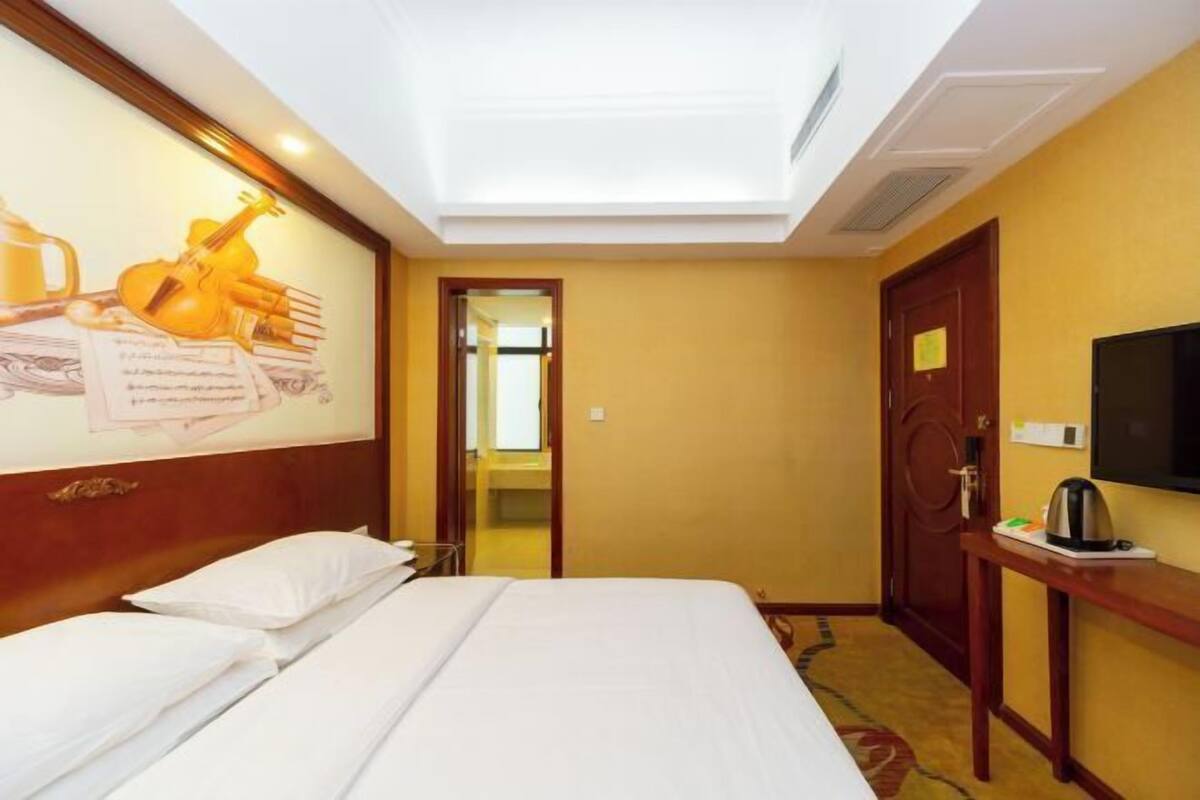 A hotel room with a bed, a wall-mounted TV, a door, and a small table with a kettle.