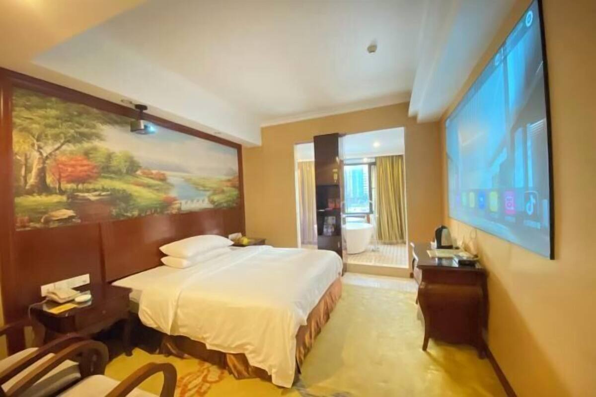 A hotel room with a bed, a desk, a chair, a large painting, and a screen displaying a landscape.