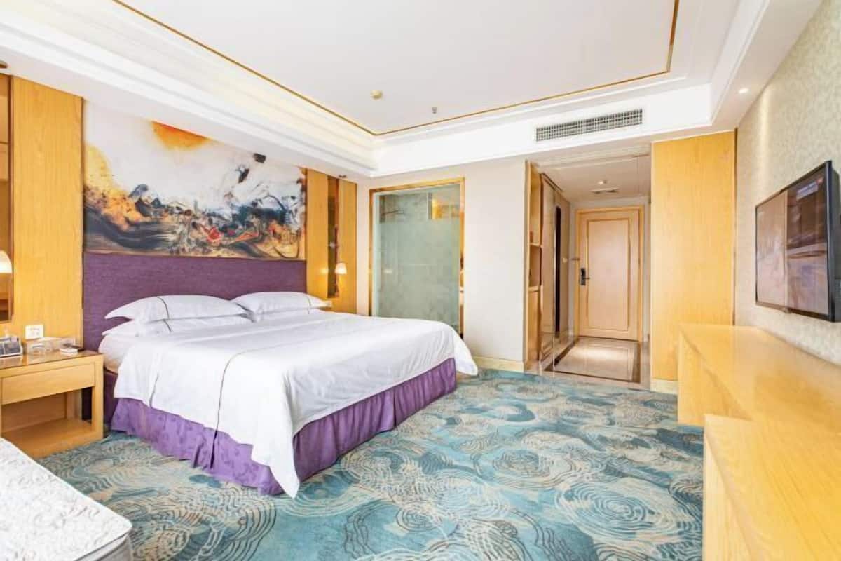 A hotel room with a large bed, a nightstand, a painting on the wall, and a door leading to another room.