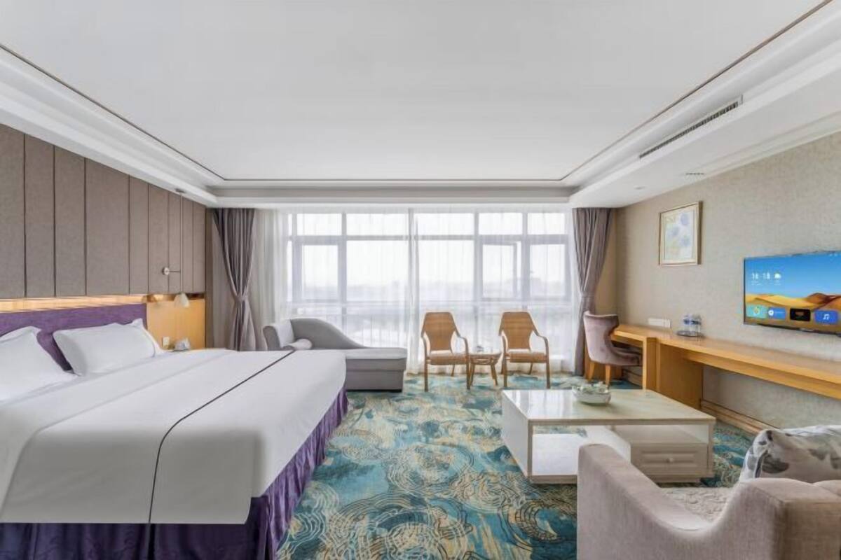 A hotel room with a large bed, a desk, two chairs, a television, and a view of the outdoors through large windows.