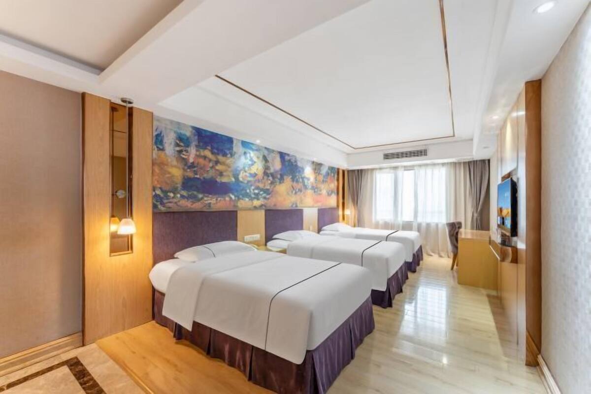 A hotel room with three beds, a large abstract painting on the wall, a flat-screen TV, and a wooden floor.