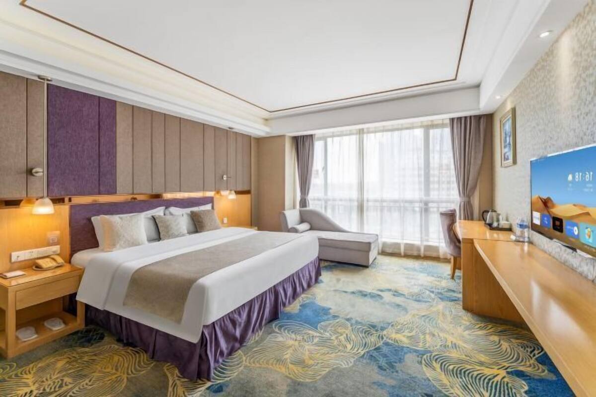 A hotel room with a large bed, a desk, a chair, and a view of the city through the window.