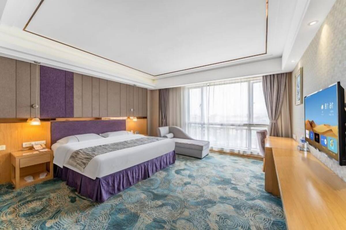 A hotel room with a bed, bedside tables, a TV, a window with curtains, and a patterned carpet.