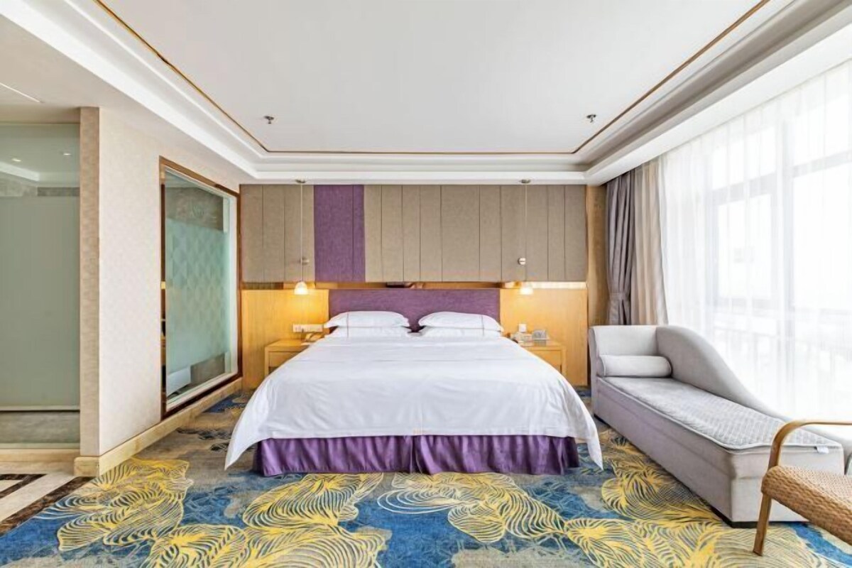 A hotel room with a large bed, a sofa, a glass shower, and a view of the city.
