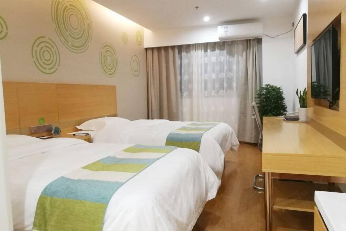 A hotel room with two beds, a wooden headboard, a desk, a chair, a potted plant, and a window with curtains.