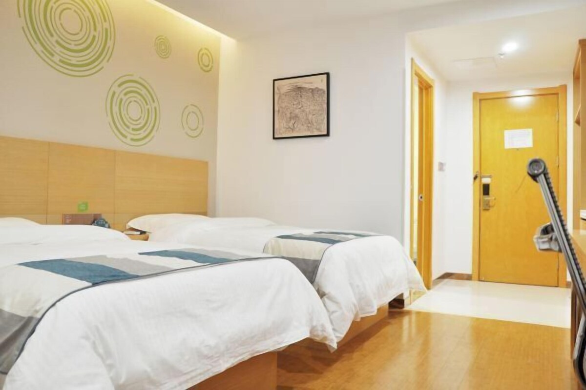 A hotel room with two beds, a wooden headboard, a framed artwork on the wall, and a door with a vacuum cleaner.