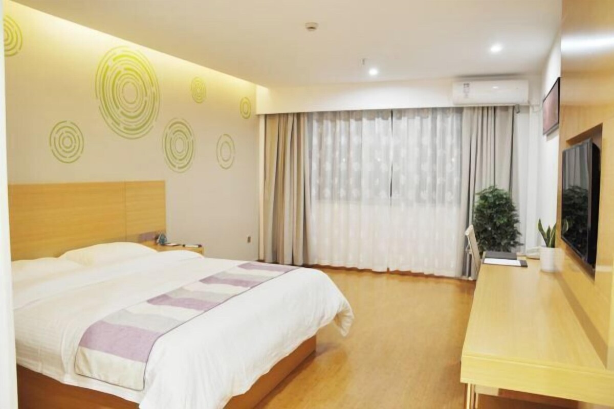 A hotel room with a bed, a desk, a potted plant, and a window with curtains.