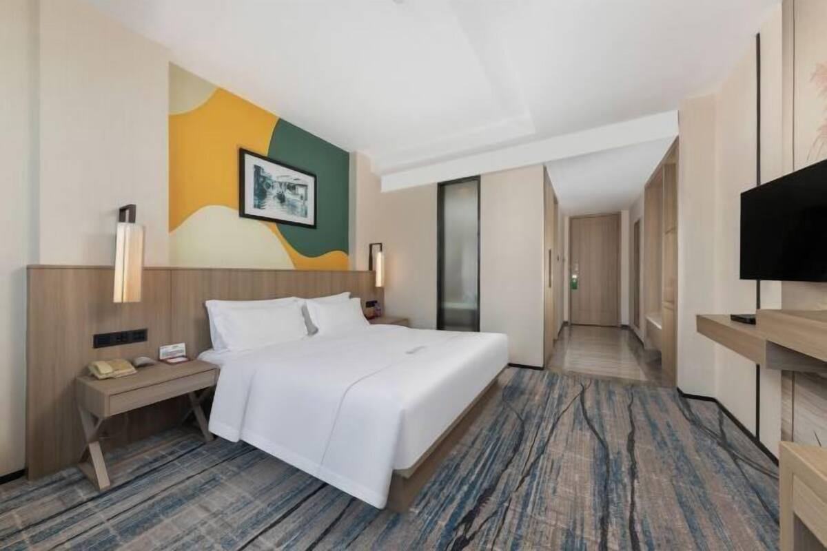 A hotel room with a bed, bedside table, lamp, artwork on the wall, and a TV.