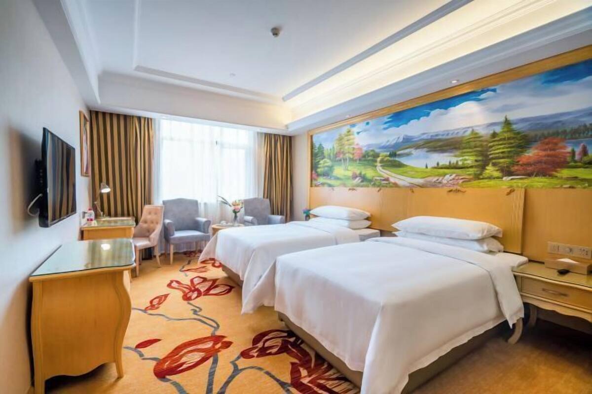 A hotel room with two beds, a desk, a chair, a TV, and a painting on the wall.