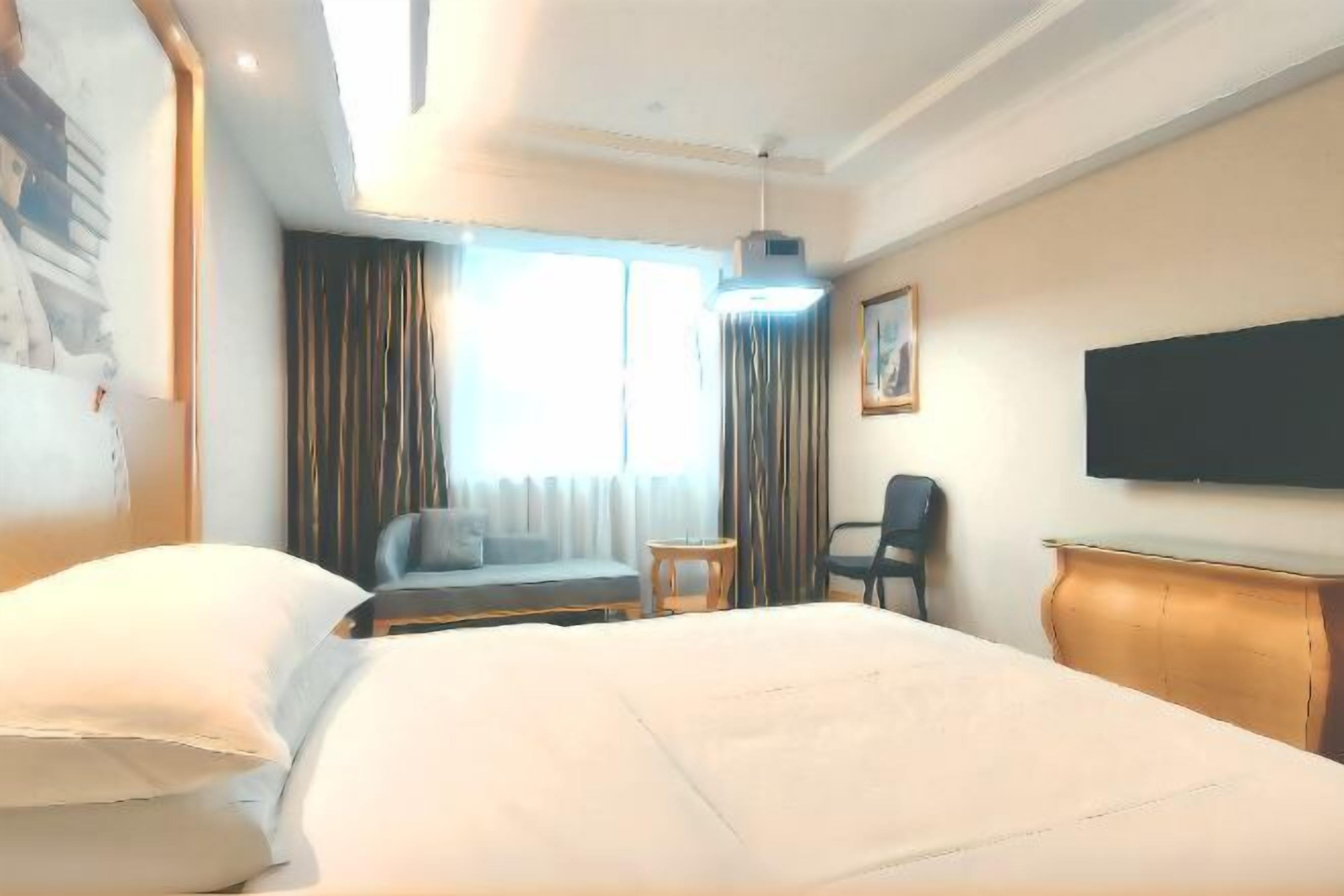 A hotel room with a large bed, a sofa, a small table, a chair, and a TV on the wall.