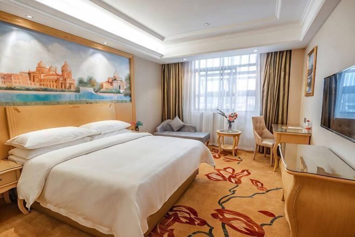A hotel room with a large bed, a desk, a chair, a small table with a vase, and a painting on the wall.